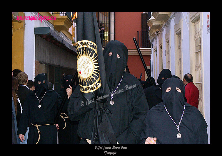 Nazareno portando la Bandera de la Compañía de Jesús de la Hermandad de Nuestra Señora de Amor y Sacrificio