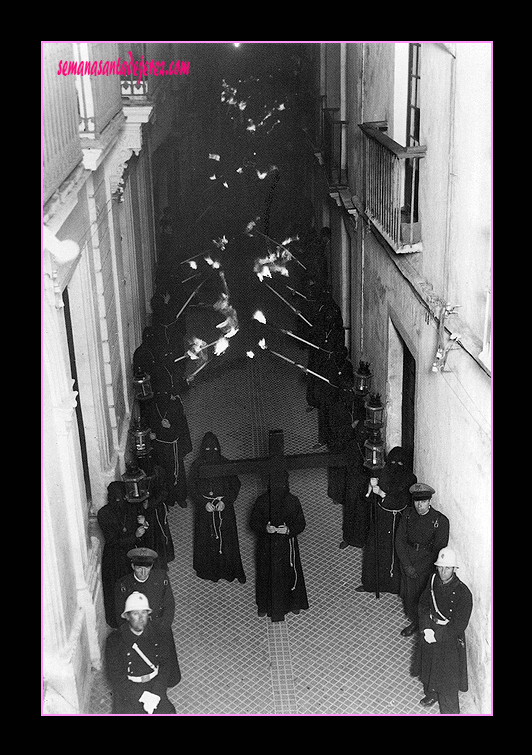 El 7 de abril de 1941 la Hermandad del Amor y Sacrificio hacía su primera Estación de Penitencia. En este momento el cortejo está en la calle Francos, casi a punto de recogerse en la Iglesia-Residencia de los Jesuitas que estuvo en la Plaza de la Compañía. Los penitentes en los primeros años no portaban cirios sino hachones (Foto: Manuel Pereiras)