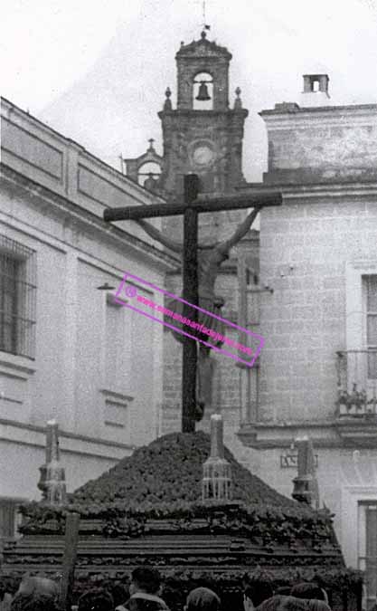 Santísimo Cristo de la Buena Muerte por la Plaza de los Angeles en el año 1959, sobre las andas prestadas del Paso de la "Chacha del Calvario" de la Hermandad del Santo Entierro. Al fondo se distingue la espadaña de la Iglesia de Santiago (Foto: Hermandad de la Buena Muerte)